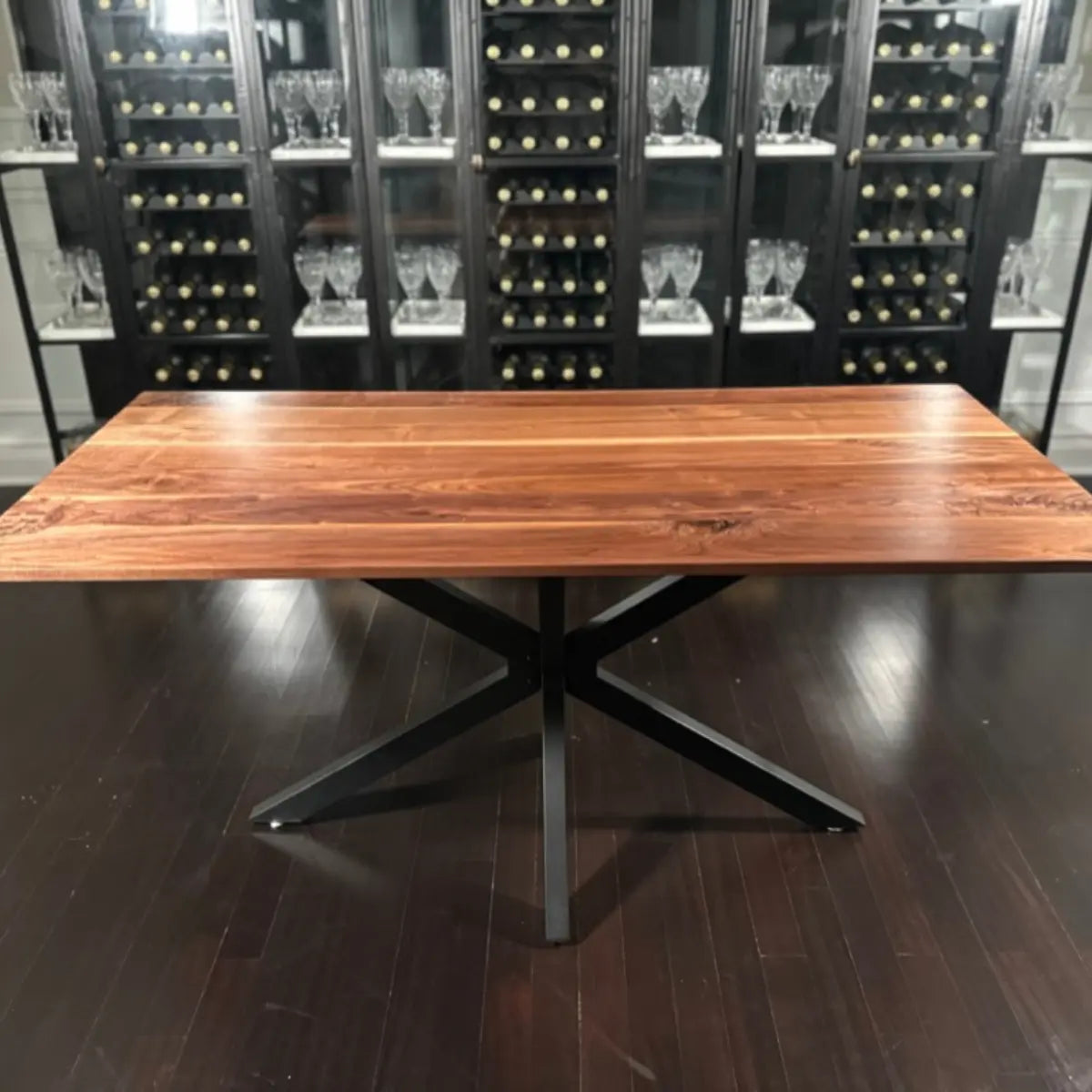 Handcrafted walnut dining table with black steel X-base, set in a modern interior with glassware display